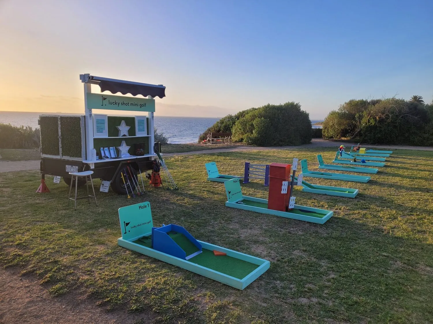 Lucky Shot Mini Golf course and branded trailer set up at a Melbourne bayside park during golden hour sunset