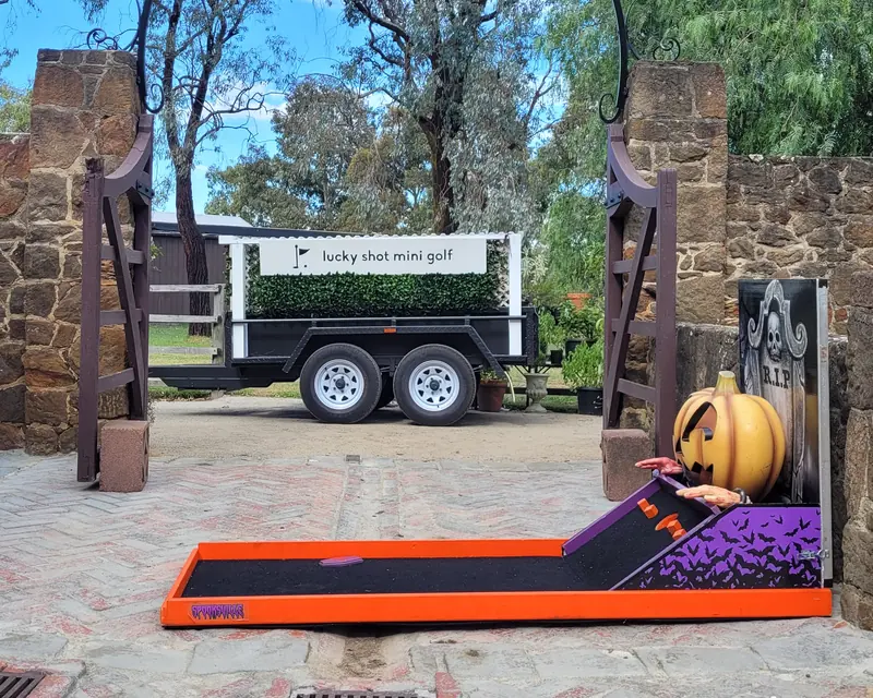 Lucky Shot's Halloween pumpkin hole set up in a bluestone courtyard, with the Lucky Shot Mini Golf trailer behind it