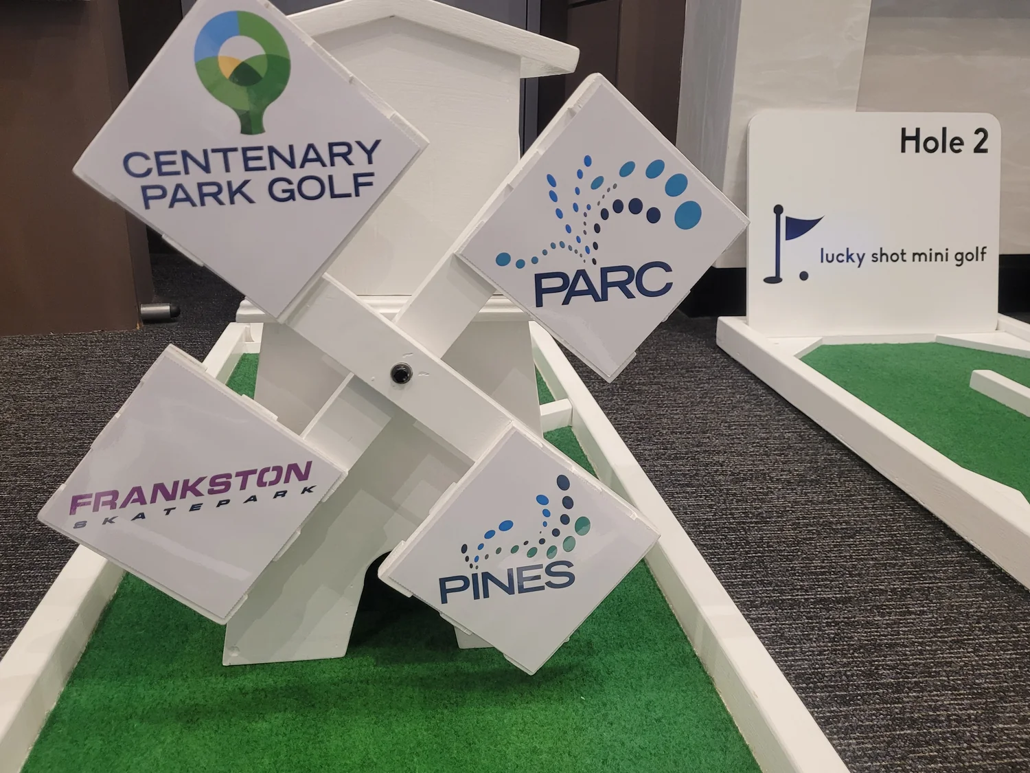 Mini golf windmill obstacle with each sail branded for a corporate client, beside a Lucky Shot hole marker
